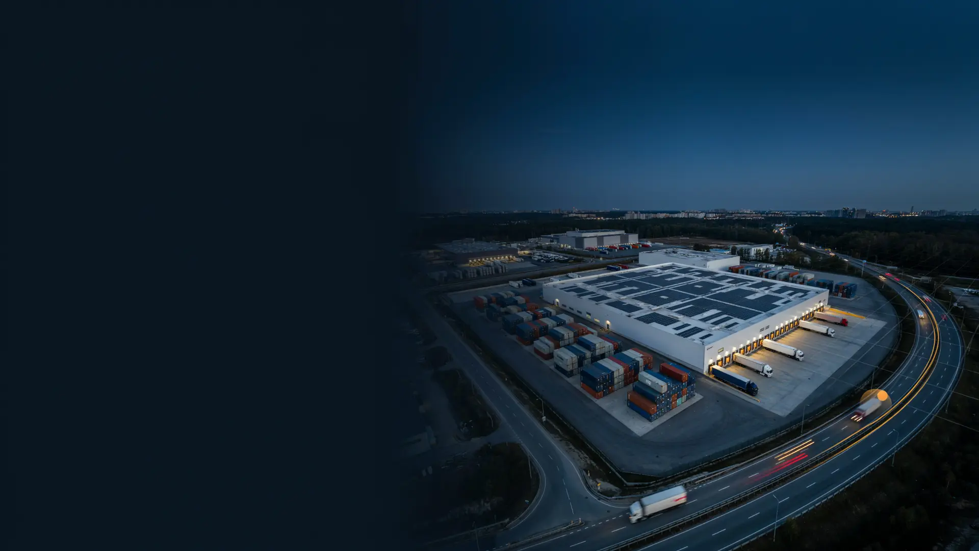 Aerial view of a distribution center at dusk with trailers at the dock doors and containers in the yard